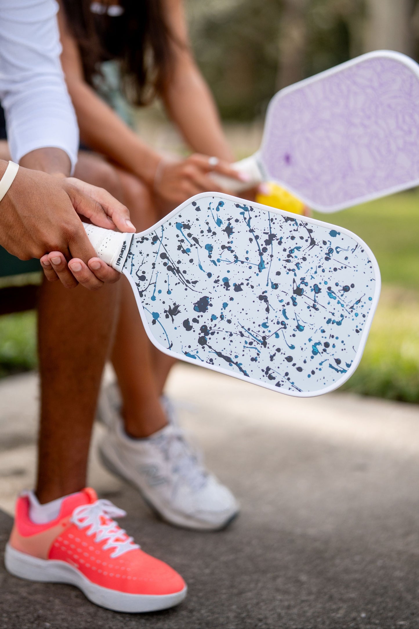 Two people holding pickleball paddles outdoors on a path.
