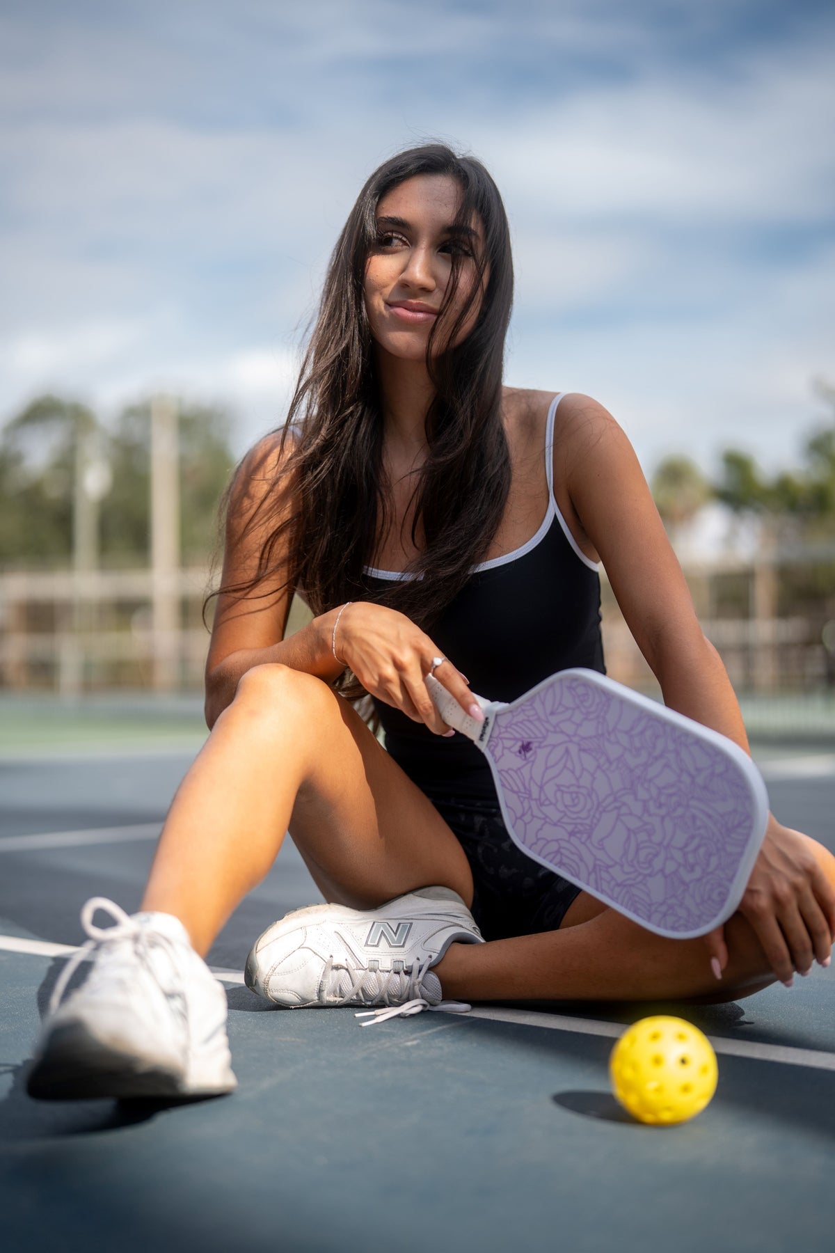 Woman sitting on a tennis court with a pink and purple Roses pickleball paddle and ball, smiling.