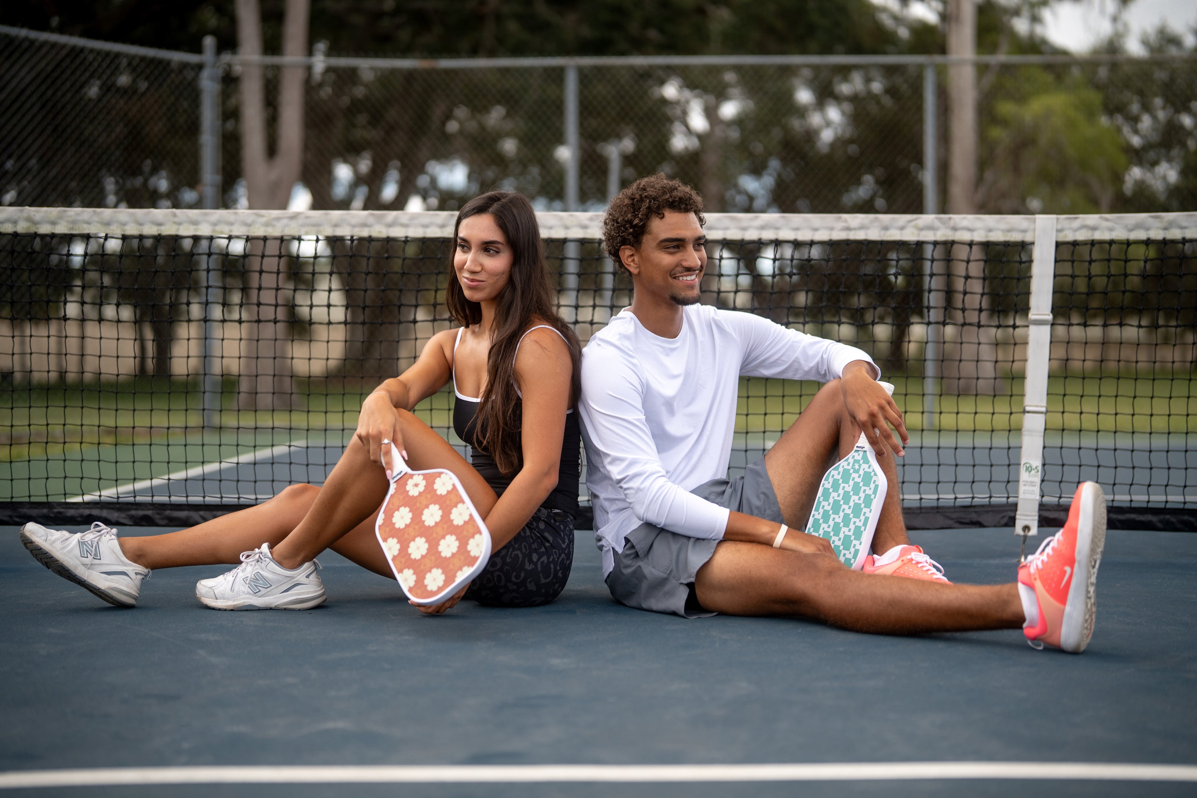 Two people sitting back to back on a pickleball court holding Red Floral and Knit green pickleball paddles, smiling.