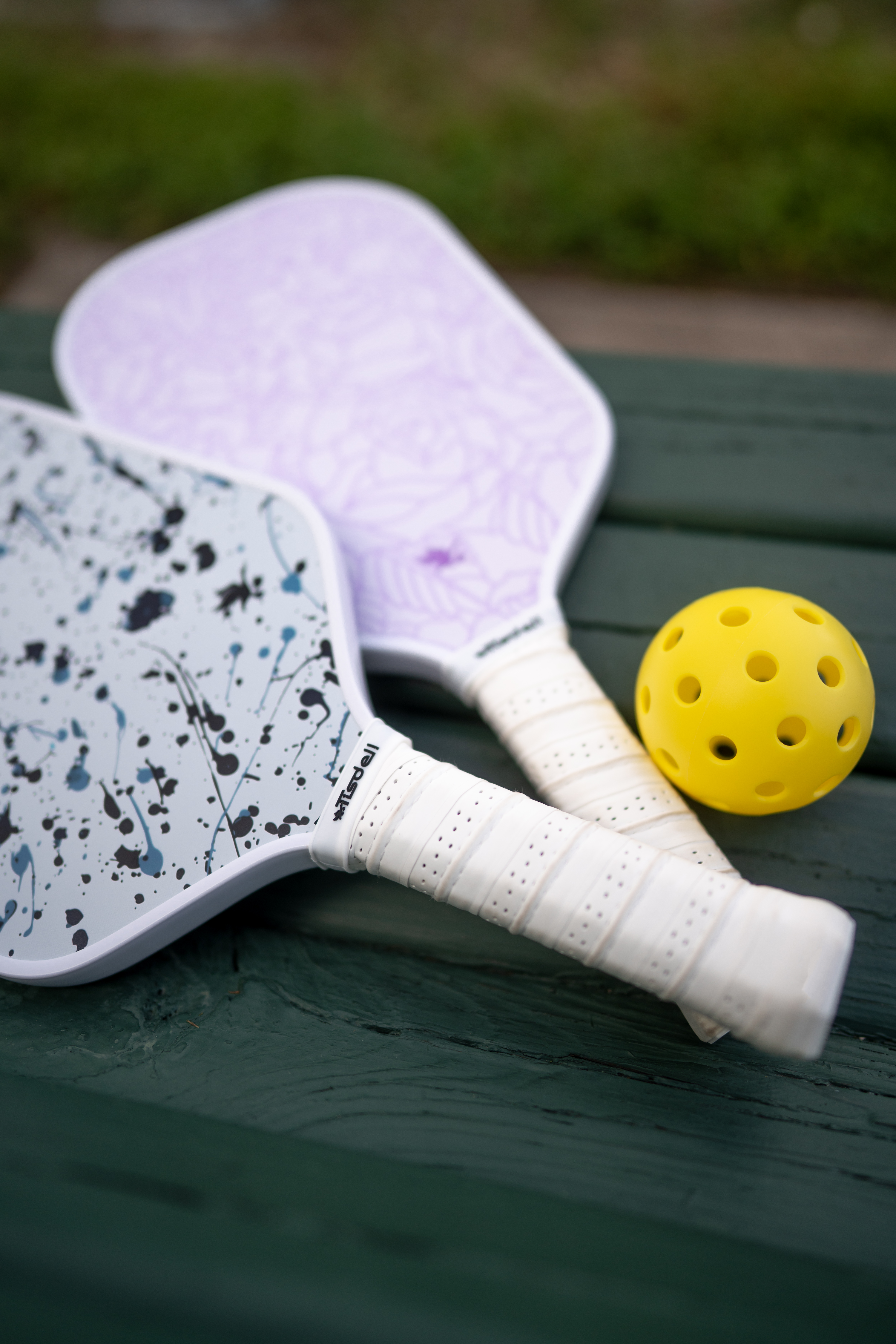 A Tisdell Everspin Series Splatter pickleball paddle and a Tisdell Everspin Series Roses pickleball paddle with a yellow ball on a green picnic table.