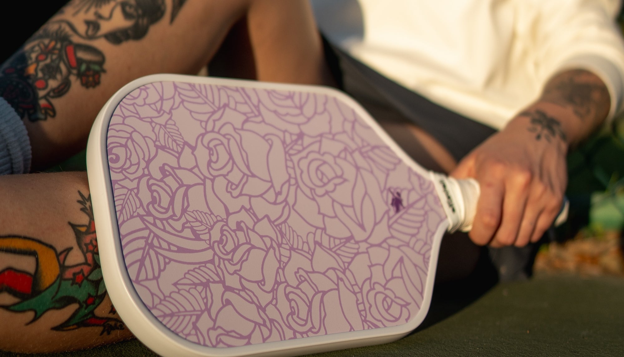 A close-up of a person holding a purple and white electric mosquito repellent fan with a floral line-art pattern, showing tattooed arms and a white shirt in an outdoor setting.