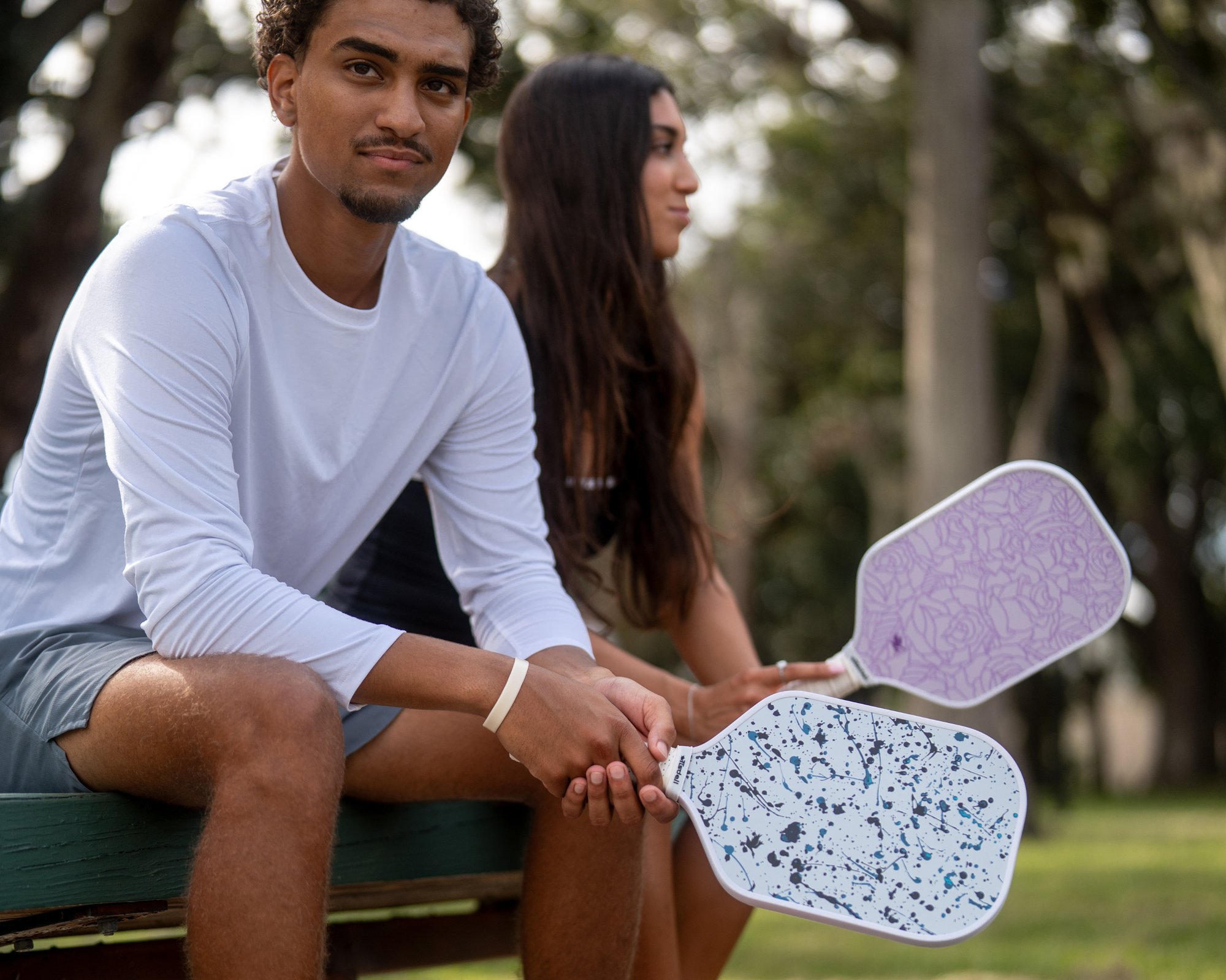 Man and woman sitting on a bench holding Tisdell pickleball paddles, one with the paint splatter design and one wit the purple / pink roses design.