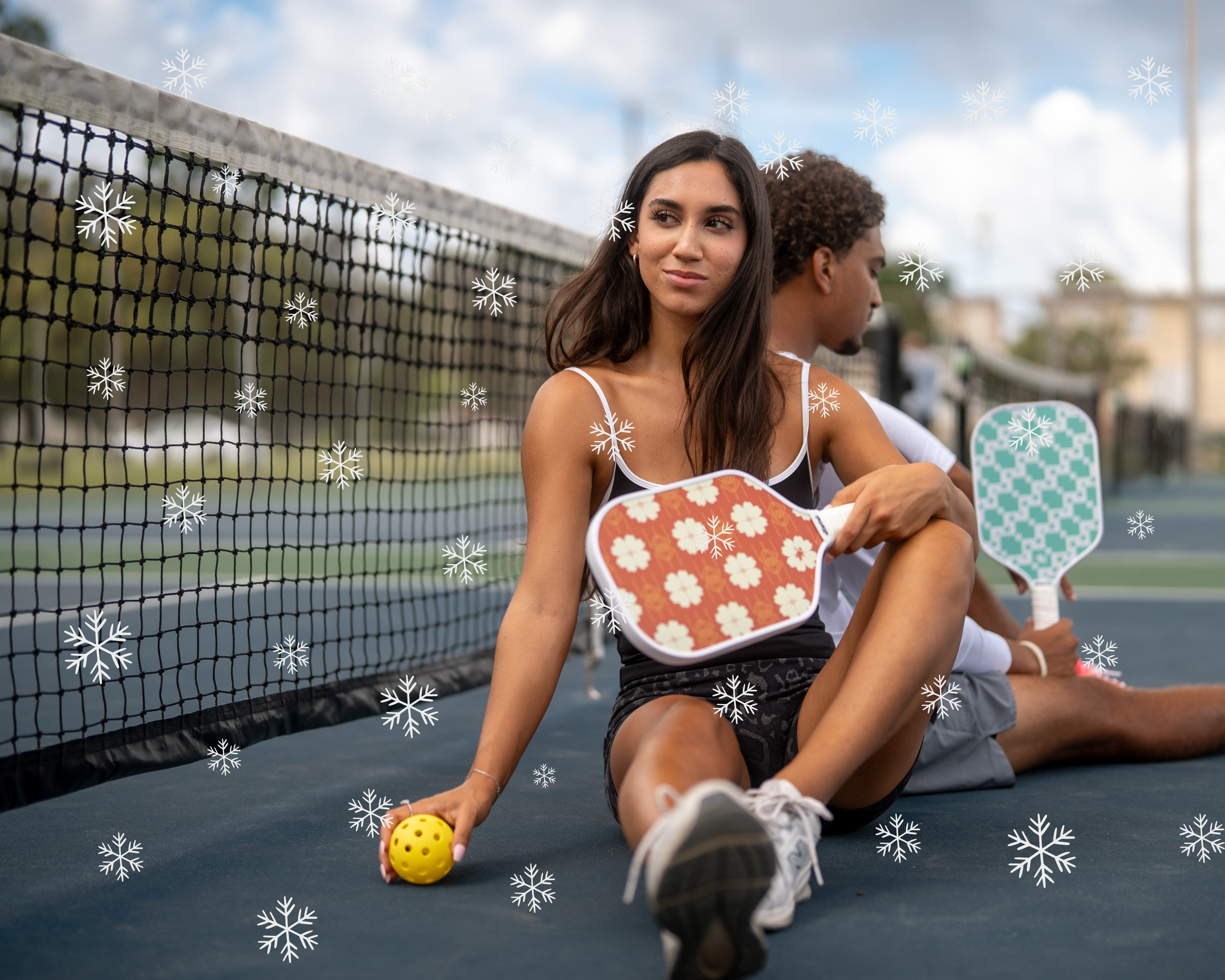 Man and Woman sitting on a pickleball court, she is holding a Tisdell Red Floral pickleball paddle and ball and he is holding a Tisdell Knit green pickleball paddle.